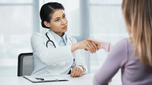 African american doctor in white coat shaking hands with patient on blurred foreground — Stock Photo