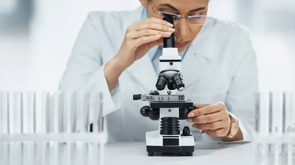 Focused african american scientist looking through microscope near test tubes in lab — Stock Photo
