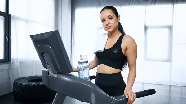 Femme sportive en sueur avec serviette sur tapis roulant regardant la caméra près de la bouteille de sport avec de l'eau dans la salle de gym — Photo de stock
