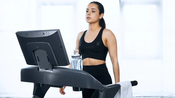 Mujer deportiva en ropa deportiva haciendo ejercicio en la cinta de correr cerca de la botella de deportes con agua en el gimnasio - foto de stock