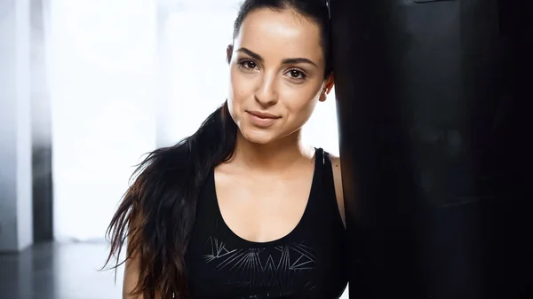 Femme sportive fatiguée regardant la caméra tout en se reposant près de sac de boxe dans la salle de gym — Photo de stock