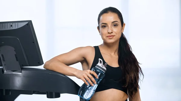 Sporty woman in sportswear holding sports bottle with water near treadmill — Stock Photo