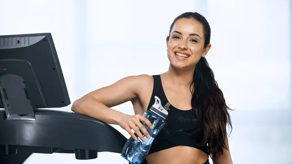 Mujer feliz en ropa deportiva celebración botella de deportes con agua cerca de la cinta de correr - foto de stock