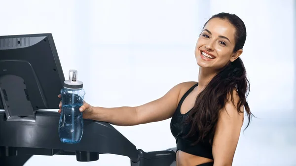 Mulher sorridente em sportswear segurando garrafa de esportes com água perto da esteira — Fotografia de Stock