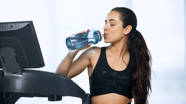 Young woman in sportswear holding sports bottle and drinking water near treadmill — Stock Photo