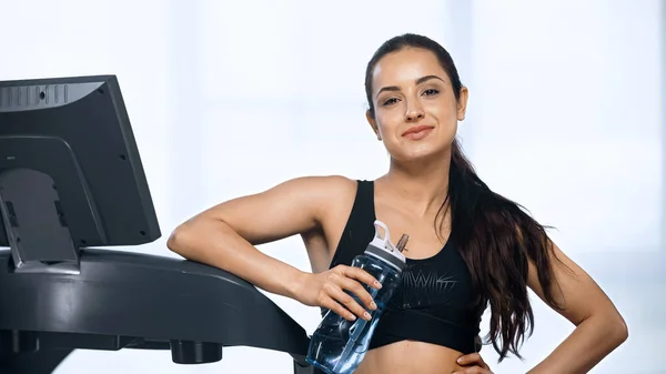 Mujer complacida en ropa deportiva sosteniendo botella deportiva con agua cerca de la cinta de correr - foto de stock