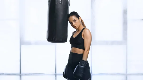 Exhausted sportswoman in boxing gloves and sportswear leaning on punching bag — Stock Photo
