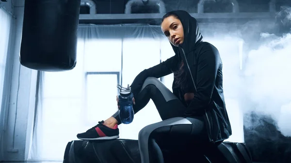 Sportliche Frau im Kapuzenpulli sitzt auf Reifen mit Sportflasche und blickt in die Kamera — Stockfoto