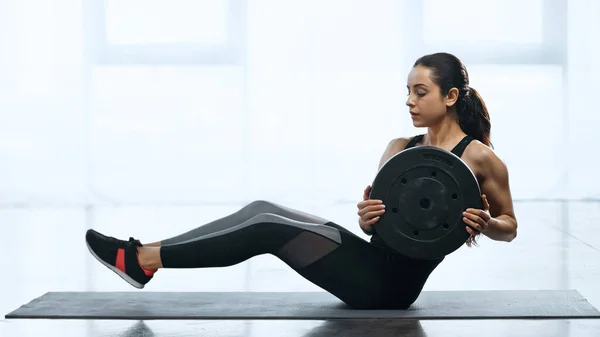 Young sportswoman doing abs with weight plate on fitness mat in gym — Stock Photo