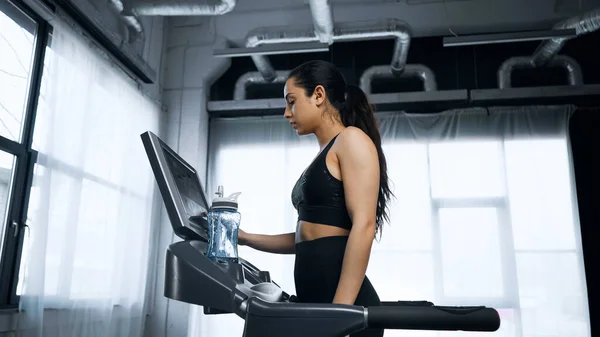 Sweaty sportswoman using touchscreen on treadmill near sports bottle with water — Stock Photo
