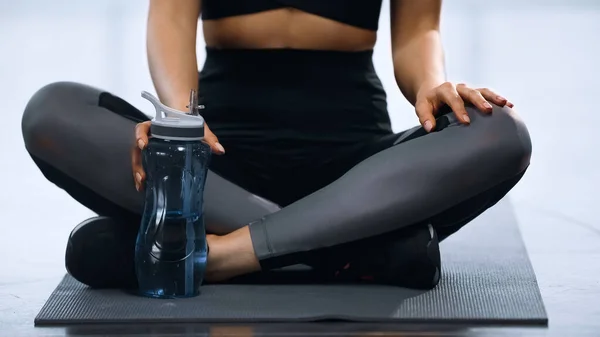 Vista recortada de la deportista sentada con las piernas cruzadas en la estera de fitness y llegar a puertos botella con agua - foto de stock