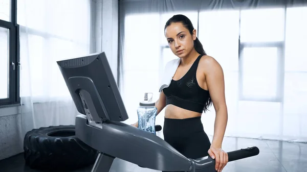 Sweaty sportswoman with towel walking on treadmill near sports bottle with water in gym — Stock Photo