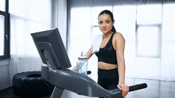 Smiling sportswoman with towel walking on treadmill near sports bottle with water in gym — Stock Photo