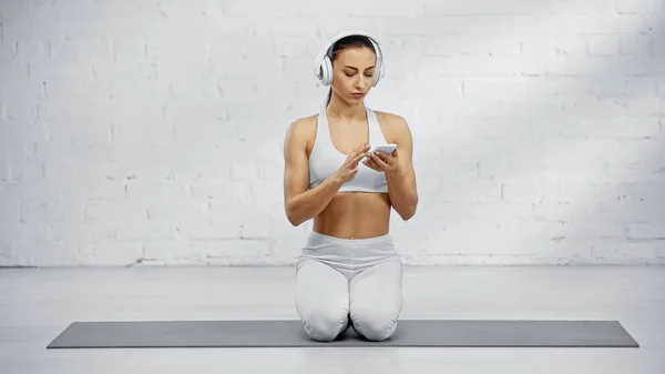 Спортсменка в наушниках с помощью смартфона на фитнес-коврике — стоковое фото