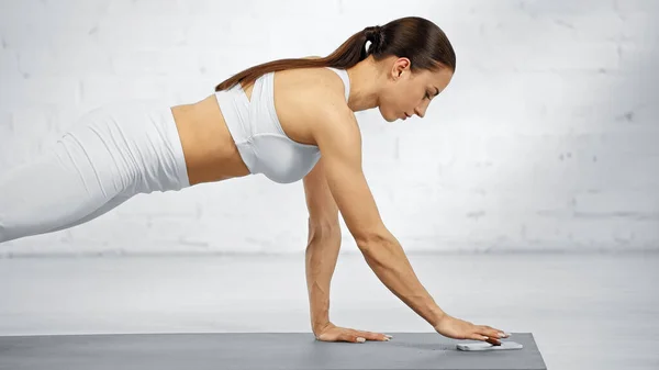 Side view of sportswoman using smartphone while standing in plank — Stock Photo