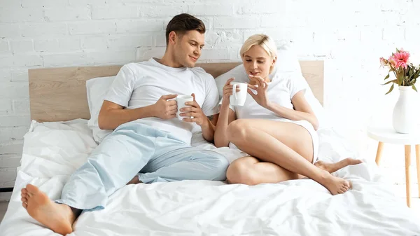 Молодая пара улыбается, держа чашки кофе в постели — стоковое фото