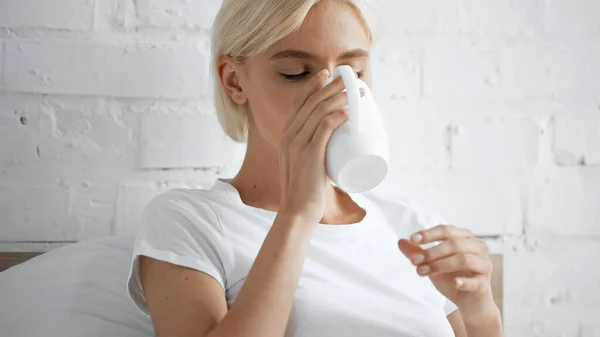 Blonde woman in white t-shirt drinking coffee in bedroom — Stock Photo