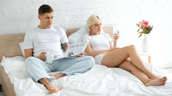 Tattooed man reading travel newspaper and holding cup of coffee near woman using smartphone in bed — Stock Photo