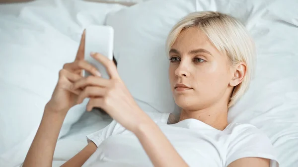 Mujer joven rubia mensajes de texto en el teléfono inteligente en el dormitorio - foto de stock