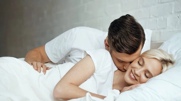 Happy man kissing smiling woman lying with closed eyes in bed — Stock Photo