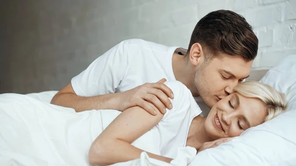 Hombre feliz besar a mujer rubia acostada con los ojos cerrados en la cama - foto de stock