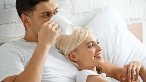 Homme tatoué buvant du café tout en étant couché avec une femme blonde heureuse au lit — Photo de stock