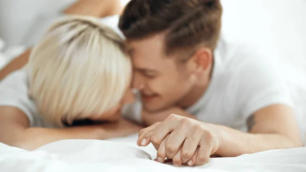 Borrosa joven pareja cogida de la mano y sonriendo en la cama - foto de stock