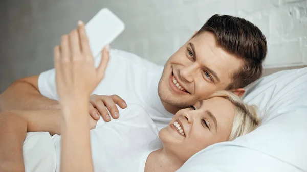 Feliz joven hombre y mujer tomando selfie en el teléfono inteligente en el dormitorio - foto de stock