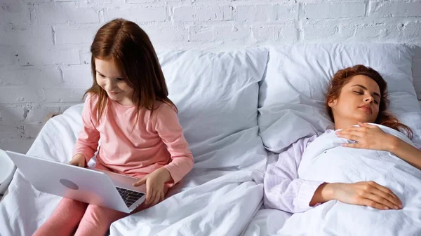 Smiling kid using laptop near mother sleeping on bed — Stock Photo