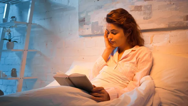 Mujer cansada leyendo libro en la cama durante la noche - foto de stock