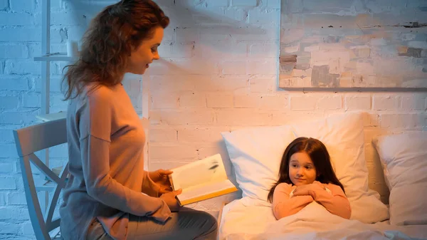 Женщина читает книгу рядом с дочерью на кровати ночью — стоковое фото