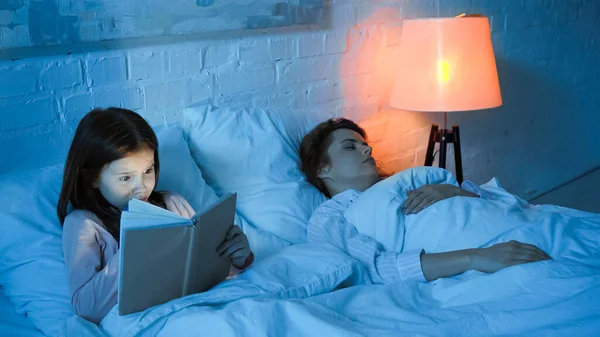 Kid leitura livro perto de mãe dormindo na cama durante a noite — Fotografia de Stock