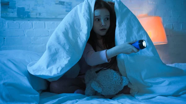 Scared kid with teddy bear holding flashlight on bed in night — Stock Photo