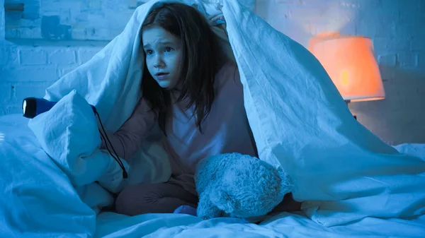 Scared child holding flashlight near teddy bear under blanket on bed — Stock Photo