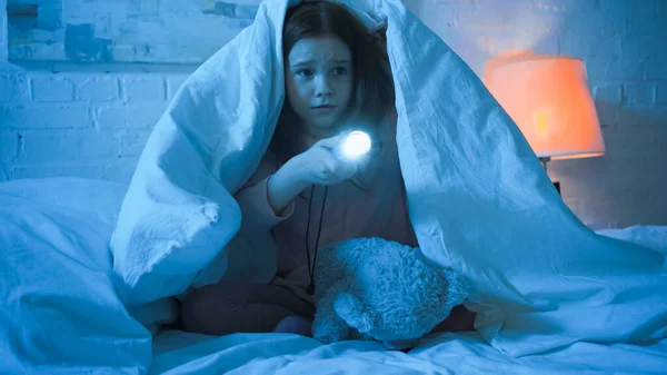 Chica sosteniendo linterna cerca de juguete suave en la cama en la noche - foto de stock