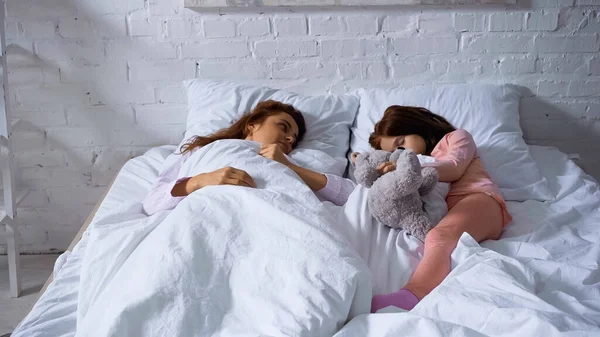 Mujer mirando a la hija dormida con oso de peluche en la cama - foto de stock