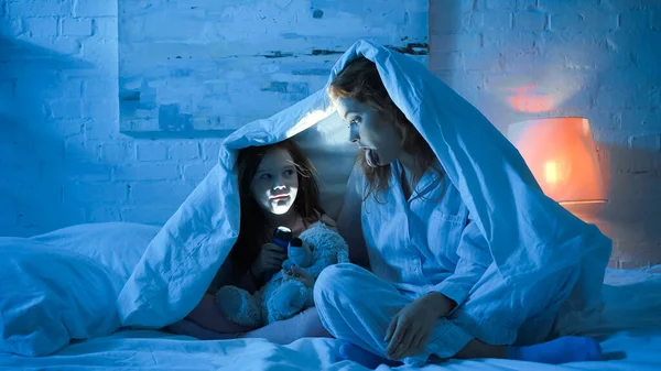 Мати стирчить язик біля дитини з м'якою іграшкою і ліхтариком на ліжку вночі — стокове фото