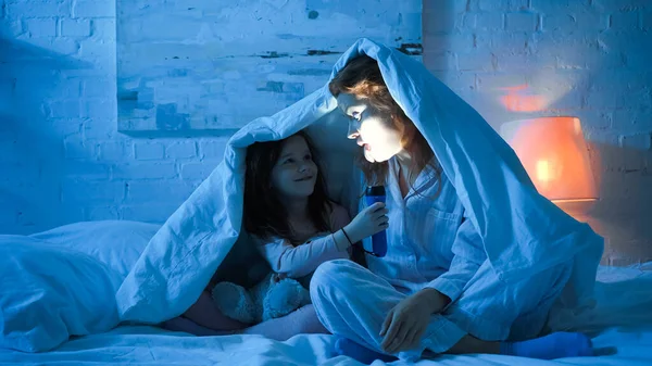 Smiling kid holding flashlight near mother sticking out tongue on bed in night — Stock Photo
