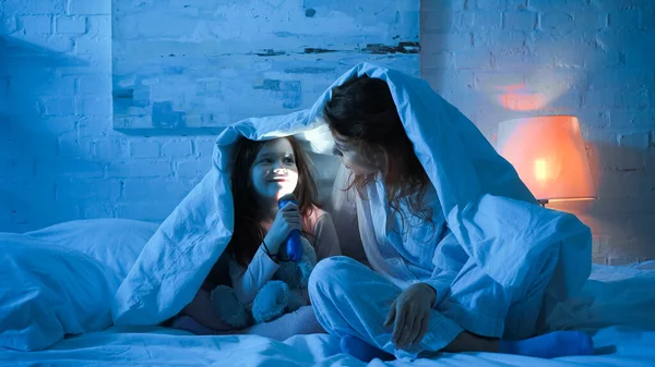 Fröhliches Kind hält Taschenlampe in der Nähe von Gesicht und Mutter auf Bett — Stockfoto