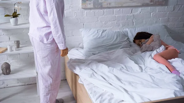 Femme en pyjama debout près d'un enfant endormi au lit le matin — Photo de stock