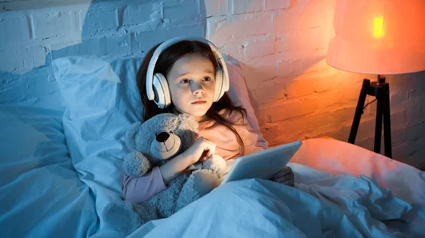 Kid in headphones holding digital tablet and teddy bear on bed — Stock Photo