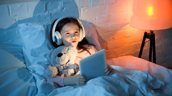 Child in headphones holding digital tablet and hugging teddy bear on bed — Stock Photo