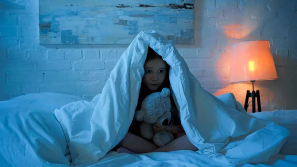 Kid holding soft toy while sitting under blanket on bed — Stock Photo