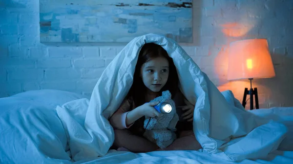 Niño con linterna y osito de peluche sentado debajo de la manta en la cama durante la noche - foto de stock