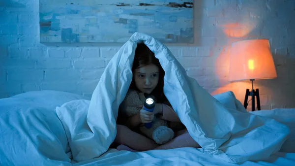 Criança assustada com brinquedo macio e ursinho sentado sob cobertor na cama — Fotografia de Stock