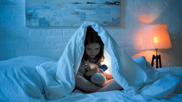 Chica asustada sosteniendo la linterna y abrazando juguete suave en la cama en la noche - foto de stock
