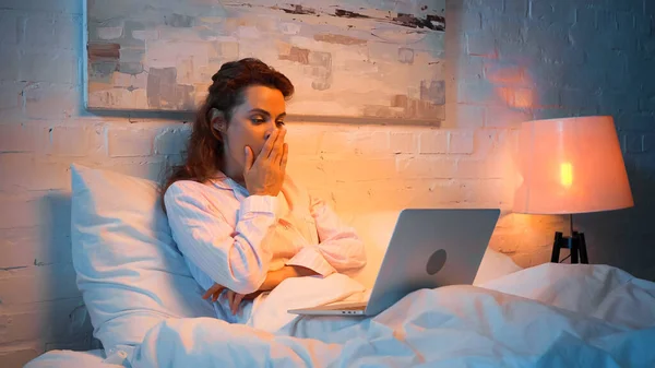 Sleepy woman in pajama looking at laptop on bed in evening — Stock Photo