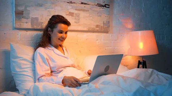 Femme gaie en pyjama en utilisant un ordinateur portable sur le lit — Photo de stock