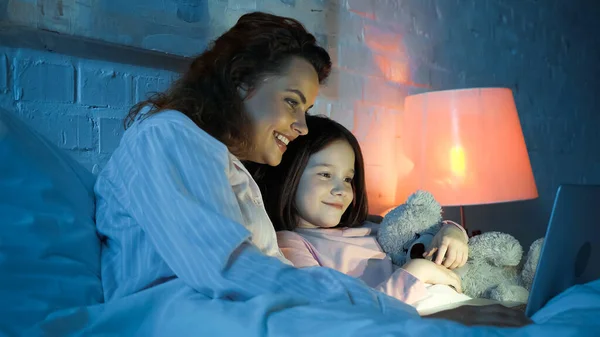 Cheerful woman using laptop near smiling daughter with soft toy on bed in night — Stock Photo