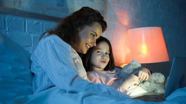 Sonriente madre e hija con osito de peluche usando portátil en la cama - foto de stock